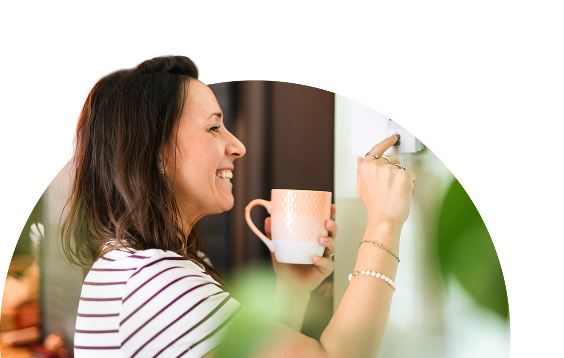 Smiling woman holding a cup of coffee while pressing buttons on a wall mounted device